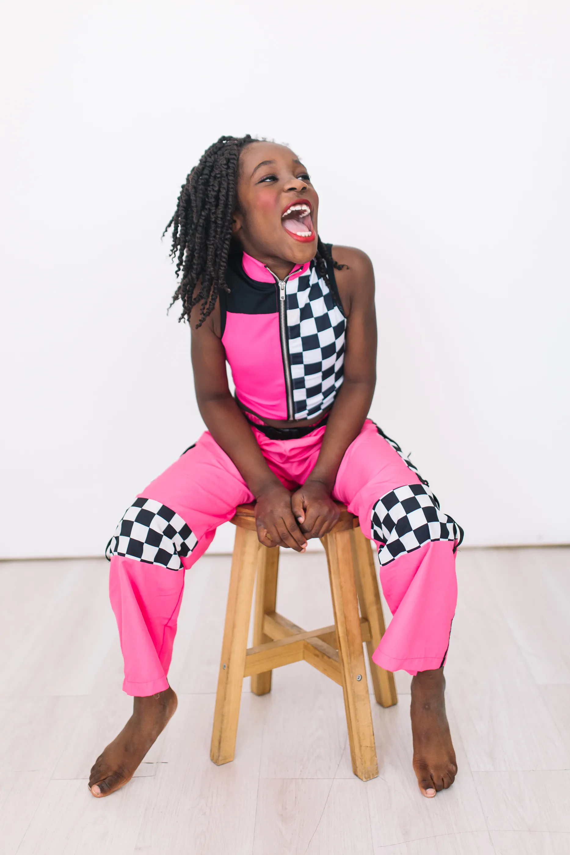 A girl mid-laugh on a wooden stool in a hot pink and checkered dance outfit.