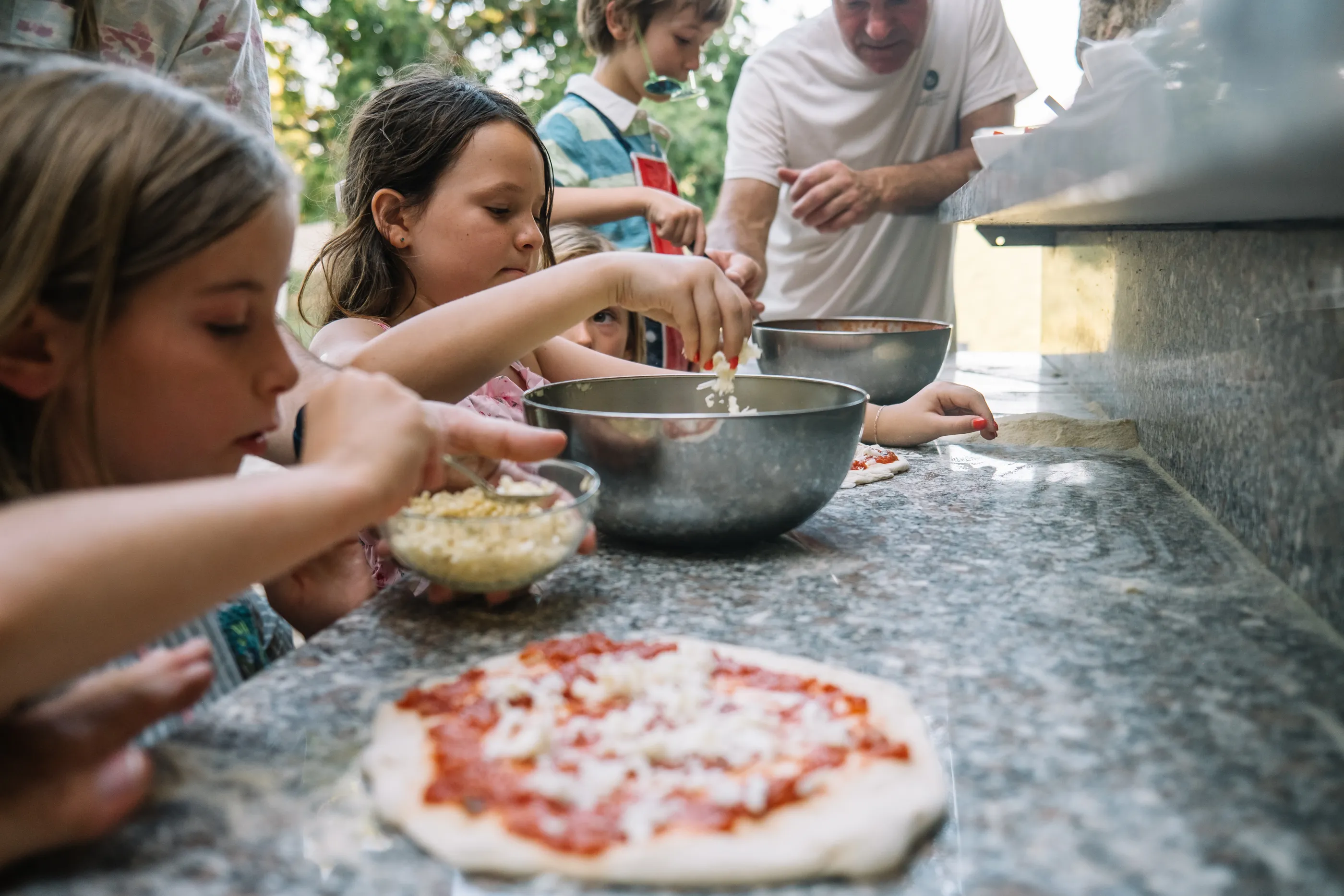 Kids around a stone counter sprinkling cheese onto a hand-stretched pizza together.